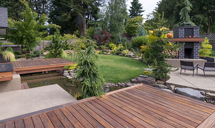 Image of backyard garden with a wooden deck, trees, and flowers.